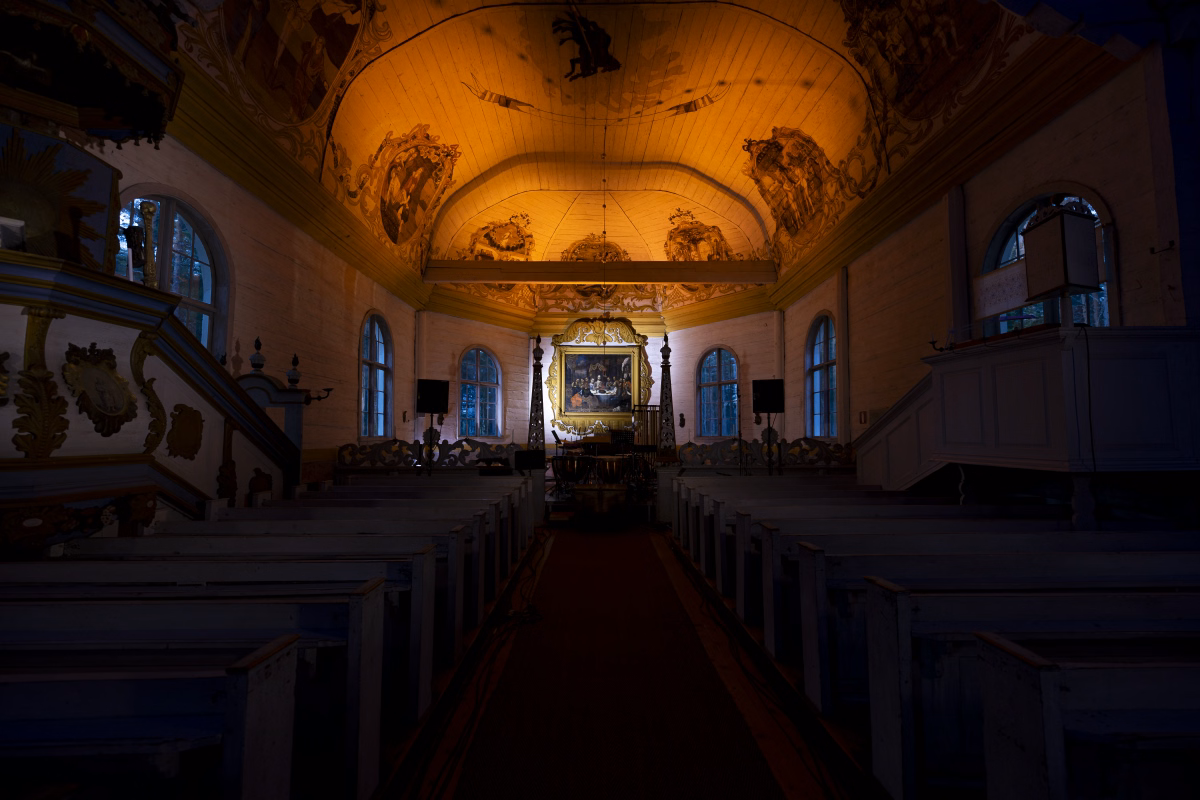 Paltaniemen kuvakirkko's nave during its live broadcast to Radio Dei, seen from the center aisle with its painted ceiling washed in warm light and the gilded altarpiece lit at the far end.