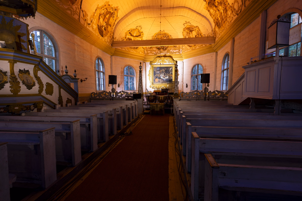 Paltaniemen kuvakirkko's nave seen from the center aisle, its painted ceiling washed in warm light and the gilded altarpiece lit at the far end.